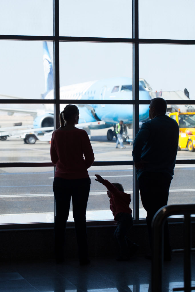 Young family watching planes at an airport standing together in front of a large glass window overlooking a jetliner being readied for takeoff